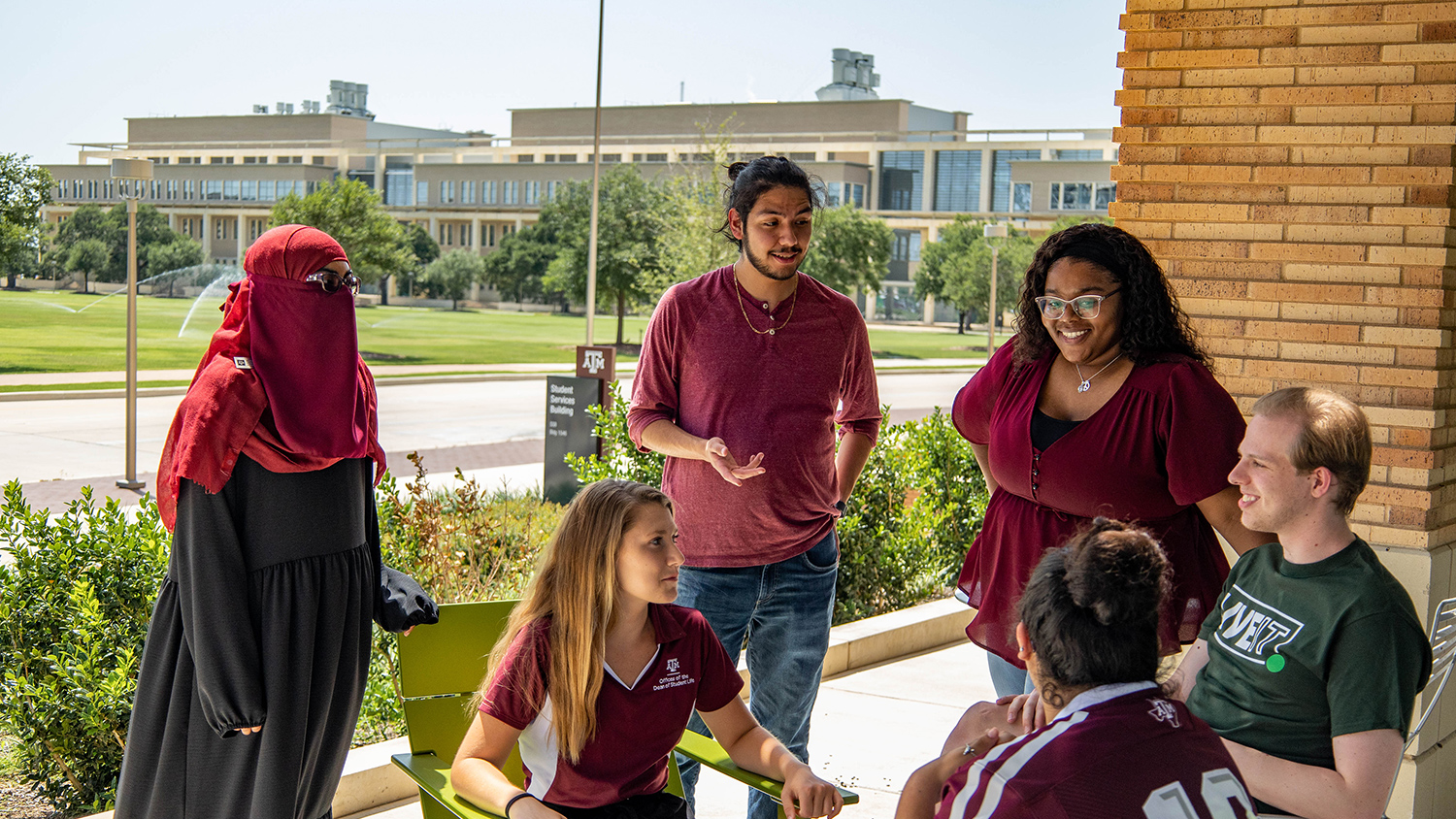 A group of six students in maroon and Green Dot apparel sitting outside on the porch of the Student Services Building, smiling and having a discussion.